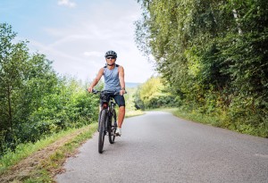 Portrait of a happy smiling man dressed in cycling clothes, helmet and sunglasses riding a bicycle on the asphalt out-of-town bicycle path. Active sporty people concept image.