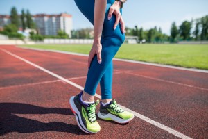Picture of a woman holding her knee and calf in pain as she stands on a jogging track.
